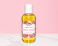 A bottle of Natural Glow Rose Gold Body Oil with a white cap, set against a solid pink background and a white marble surface.