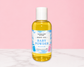 A bottle of Natural Glow For & Baby Baby Powder Body Oil, filled with yellow liquid and featuring a white cap, set against a pink background with a white marble surface.