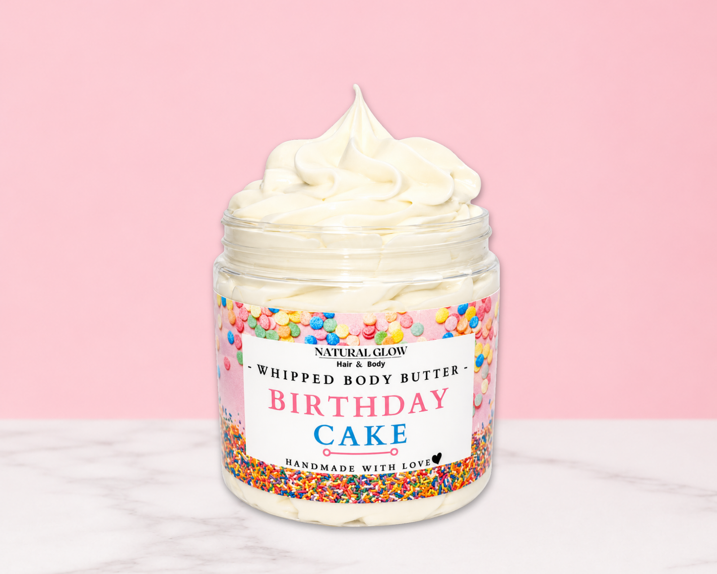 Natural Glow Birthday Cake whipped body butter in a jar with cream-colored butter swirl, pink background, and white marble surface