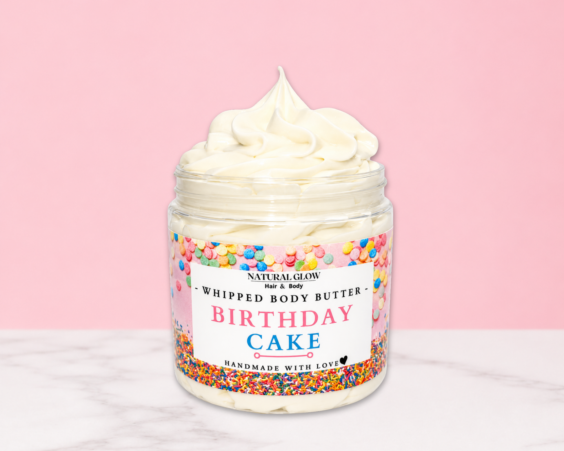 Natural Glow Birthday Cake whipped body butter in a jar with cream-colored butter swirl, pink background, and white marble surface