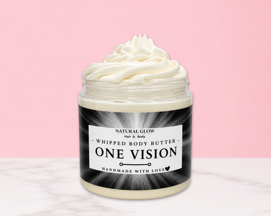 A jar of whipped body butter with a swirl of creamy white product visible at the top, set on a light marble surface against a solid pink background. The jar has a black and white label reading 'ONE VISION NATURAL GLOW Whipped Body Butter Handmade with Love'.