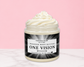 A jar of whipped body butter with a swirl of creamy white product visible at the top, set on a light marble surface against a solid pink background. The jar has a black and white label reading 'ONE VISION NATURAL GLOW Whipped Body Butter Handmade with Love'.