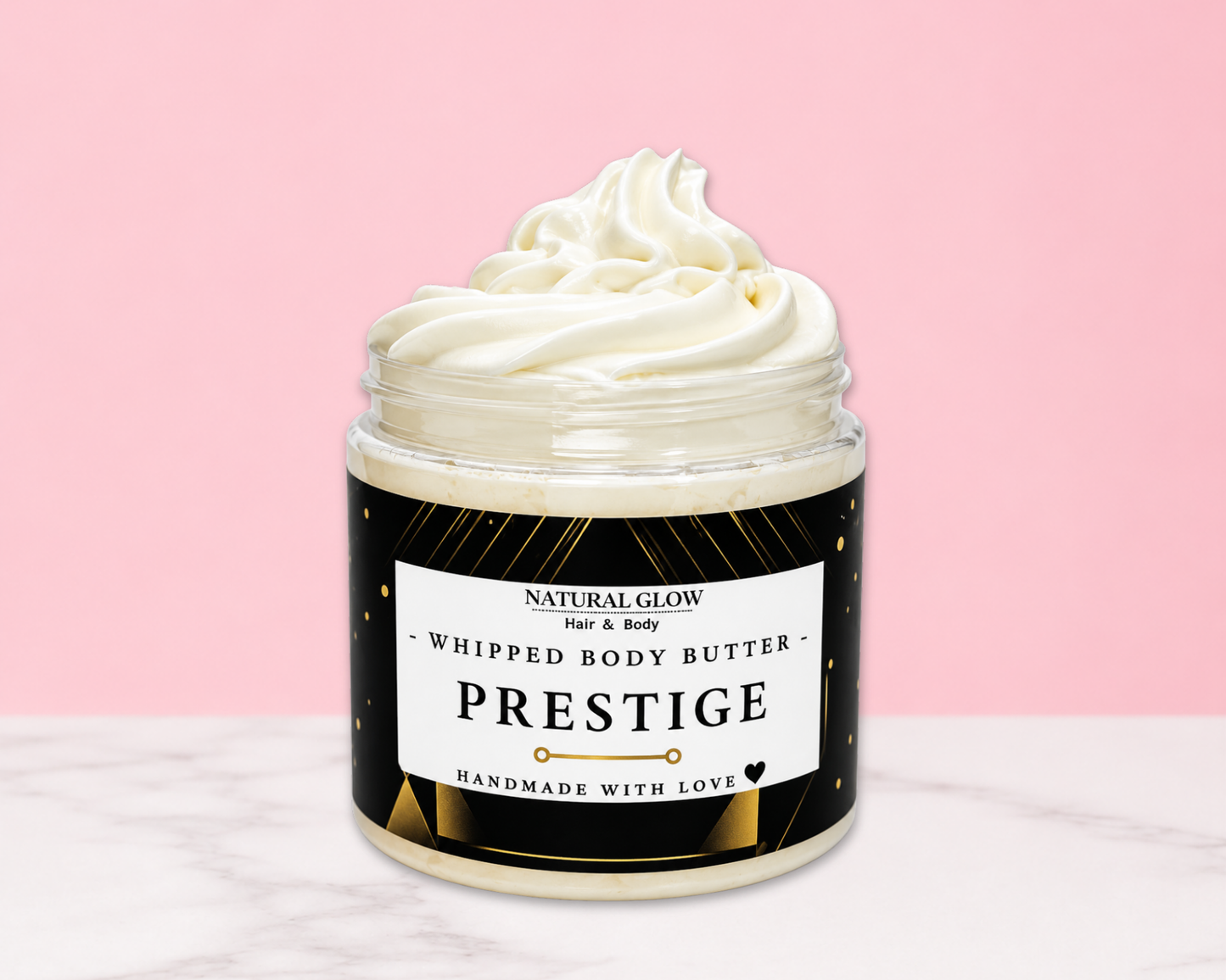 A jar of whipped body butter with a swirl of creamy white product visible at the top, set against a pink background with a white marble surface. The label reads 'NATURAL GLOW Just & Body PRESTIGE HANDMADE WITH LOVE'.
