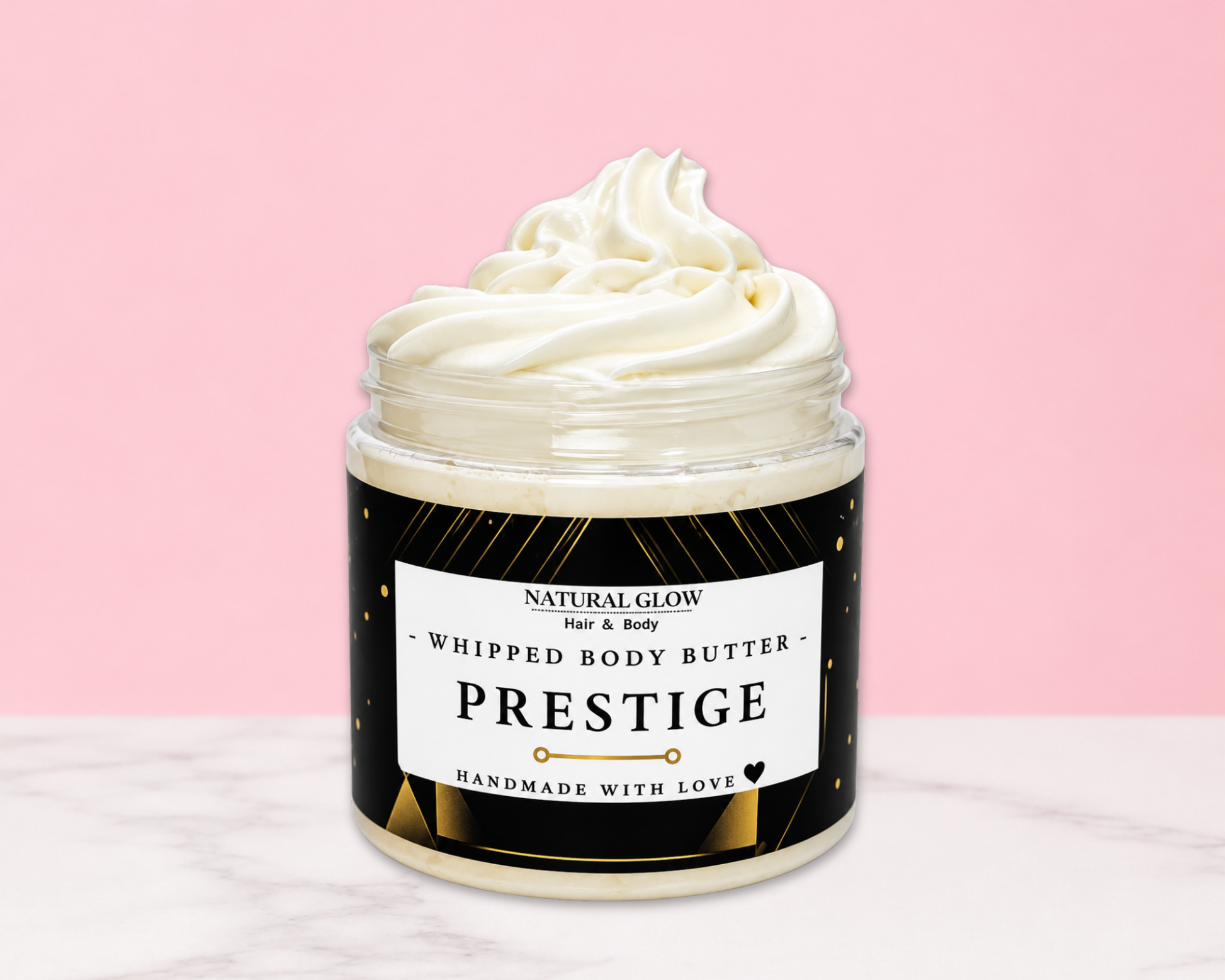 A jar of whipped body butter with a swirl of creamy white product visible at the top, set against a pink background with a white marble surface. The label reads 'NATURAL GLOW Just & Body PRESTIGE HANDMADE WITH LOVE'.