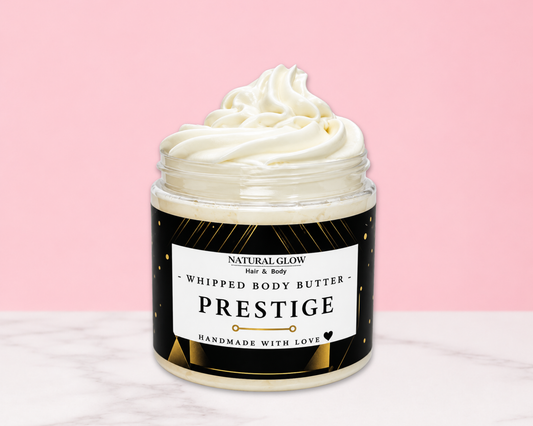 A jar of whipped body butter with a swirl of creamy white product visible at the top, set against a pink background with a white marble surface. The label reads 'NATURAL GLOW Just & Body PRESTIGE HANDMADE WITH LOVE'.