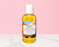 A bottle of Natural Glow Body Oil in Pumpkin Spice scent, filled with yellow oil and featuring a white cap, set against a pink and white background.